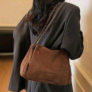 May include: A brown suede shoulder bag with a chain strap. The bag has a rectangular shape with a soft, textured appearance. The bag is worn over a dark blazer.