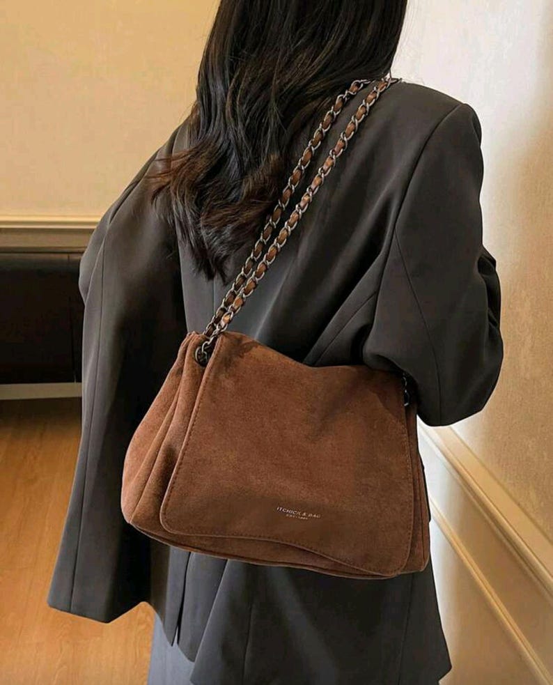 May include: A brown suede shoulder bag with a chain strap. The bag has a rectangular shape with a soft, textured appearance. The bag is worn over a dark blazer.