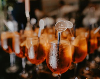 May include: A collection of stemmed glasses filled with an orange-colored beverage, likely a cocktail. Each glass has a straw and a small, round sign with text. The drinks are arranged on a dark surface, suggesting a celebratory event.