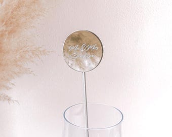 May include: A silver-colored drink stirrer with a round, reflective top. The top is engraved with the words "Mr & Mrs Sanderson" in an elegant script. The stirrer is placed in a clear glass, with a soft pink background.