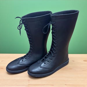 May include: A pair of black, knee-high boots with a lace-up design. The boots have a rounded toe and a flat sole. The boots are made of a smooth material and are set against a green background.