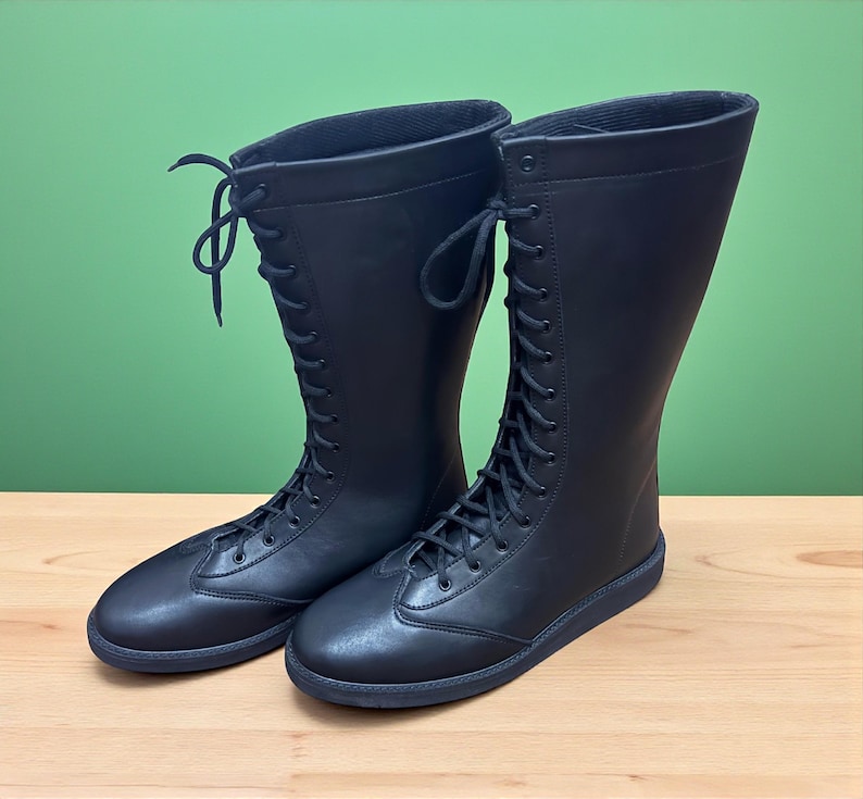 May include: A pair of black, knee-high boots with a lace-up design. The boots have a rounded toe and a flat sole. The boots are made of a smooth material and are set against a green background.