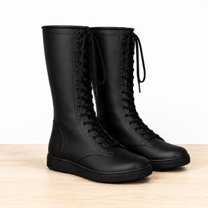 May include: A pair of matte black knee-high boots featuring a smooth finish, a rounded toe, and a full-length front lace-up closure with black eyelets and matching black laces. The boots have a flat, solid black rubber sole and a clean, seamless design.