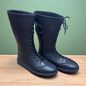 May include: A pair of black, knee-high boots with a lace-up design. The boots have a flat sole and a smooth, matte finish. The laces extend up the front of the boots, and the top of the boots are slightly flared.