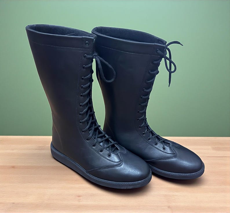 May include: A pair of black, knee-high boots with a lace-up design. The boots have a flat sole and a smooth, matte finish. The laces extend up the front of the boots, and the top of the boots are slightly flared.