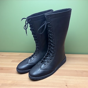 May include: A pair of black, knee-high boots with a lace-up design. The boots are made of a smooth material and have a rounded toe. The boots are displayed on a wooden surface with a green background.