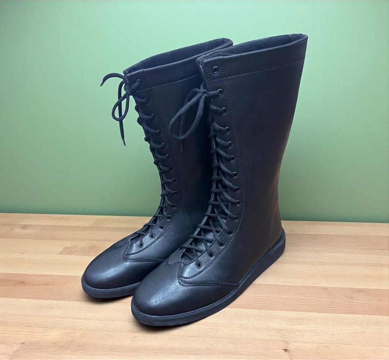 May include: A pair of black, knee-high boots with a lace-up design. The boots are made of a smooth material and have a rounded toe. The boots are displayed on a wooden surface with a green background.