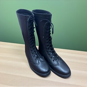 May include: A pair of tall, black leather riding boots with a lace-up design. The boots have a smooth finish and are displayed on a light-colored wooden surface against a green background. The boots appear to be new and unworn.
