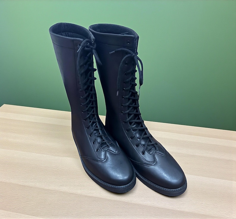 May include: A pair of tall, black leather riding boots with a lace-up design. The boots have a smooth finish and are displayed on a light-colored wooden surface against a green background. The boots appear to be new and unworn.