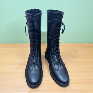 May include: A pair of black, knee-high boots with a lace-up design. The boots are made of a smooth material and have a rounded toe. The laces extend up the front of the boots. The boots are displayed on a wooden surface.