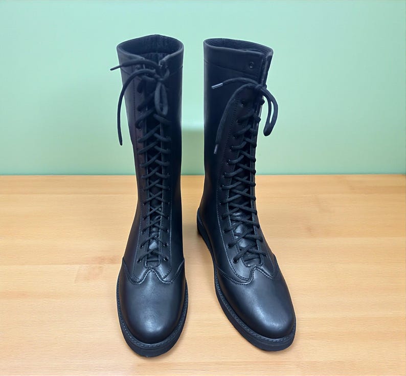 May include: A pair of black, knee-high boots with a lace-up design. The boots are made of a smooth material and have a rounded toe. The laces extend up the front of the boots. The boots are displayed on a wooden surface.