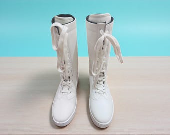 Pode incluir: Um par de botas brancas até o joelho com cadarços brancos. As botas têm bico redondo e sola branca. O fundo é uma parede azul clara e uma superfície de madeira clara.