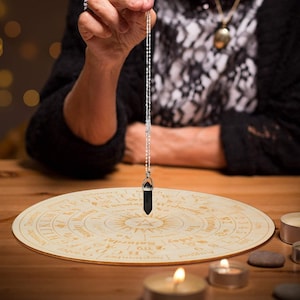May include: A black crystal pendulum suspended over a wooden divination board. The board is round with engraved text and symbols. The pendulum hangs from a silver chain. Candles are lit around the board, creating a mystical atmosphere.