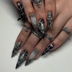 May include: Close-up of hands adorned with elaborate, long, pointed nail art in black, silver, and white. The nails feature intricate designs, including ornate silver embellishments, pearls, and studs. Several silver rings with detailed designs complement the nail art.