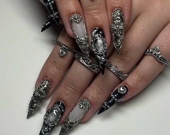 May include: Close-up of hands adorned with elaborate, long, pointed nail art in black, silver, and white. The nails feature intricate designs, including ornate silver embellishments, pearls, and studs. Several silver rings with detailed designs complement the nail art.