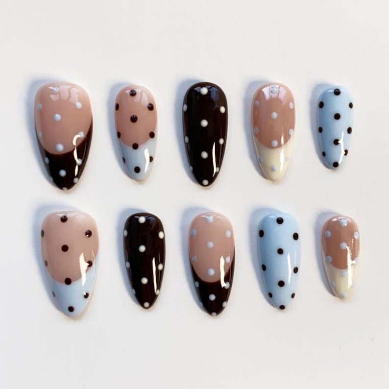 May include: A collection of ten oval false nails. The designs include solid dark brown, light blue with white polka dots, and a two-tone design with a light brown top and dark brown bottom. Each nail is approximately 2 cm long.