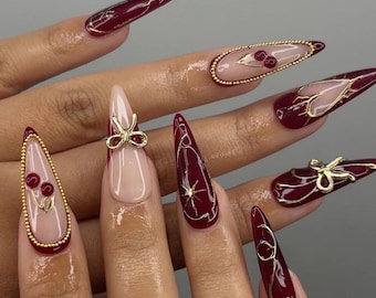 May include: Long, almond-shaped burgundy nails with gold accents. Some nails feature gold bow charms, while others have gold line designs. One nail is nude with a gold bow. Some nails have gold beaded borders and cherry designs.