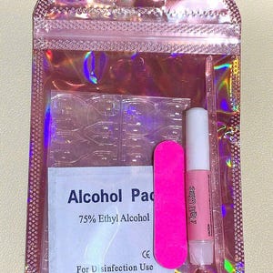 May include: A pink holographic zip-lock bag containing nail supplies. Inside are clear nail tips, a pink emery board, a tube of nail glue, and an alcohol pad with the text "Alcohol Pad 75% Ethyl Alcohol For Disinfection Use".