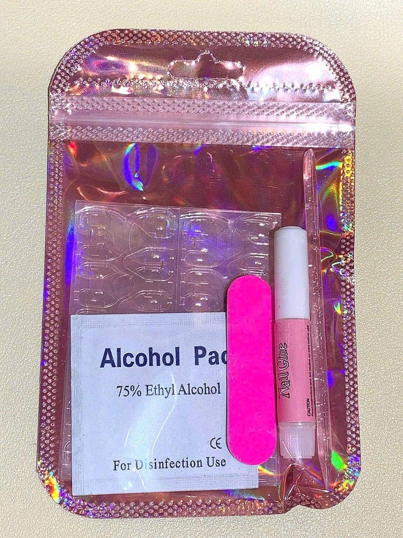 May include: A pink holographic zip-lock bag containing nail supplies. Inside are clear nail tips, a pink emery board, a tube of nail glue, and an alcohol pad with the text "Alcohol Pad 75% Ethyl Alcohol For Disinfection Use".