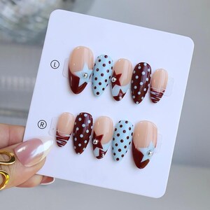 May include: A set of almond-shaped false nails in various designs. The nails feature a nude base with accents in burgundy, light blue with white polka dots, and a star design with a rhinestone. The nails are displayed on a white card.
