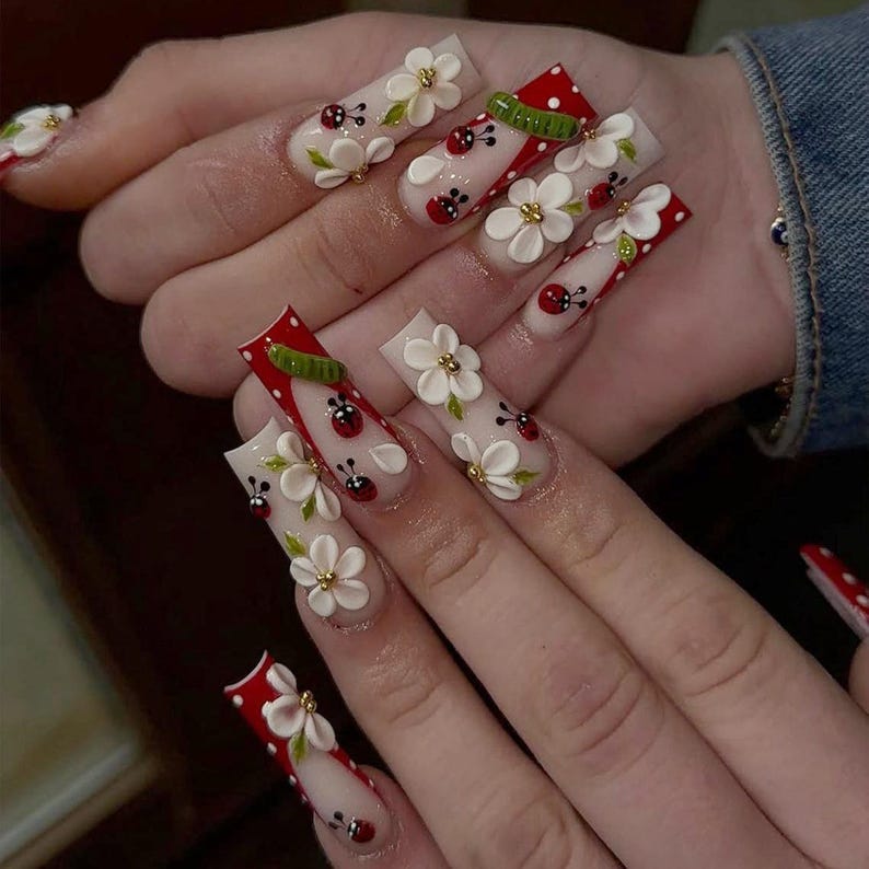 May include: Long, square-shaped artificial nails with a clear base, adorned with white flowers, gold accents, ladybugs, and red polka dot designs. Some nails feature a red and green design. The nails are on a hand.