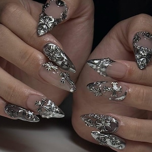 May include: Close-up of a set of long, pointed fingernails with a clear base and silver embellishments. The silver designs include crosses, floral patterns, and ornate metal accents, creating a gothic or edgy aesthetic.
