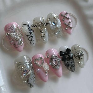May include: A set of ten almond-shaped artificial nails. The nails are painted in shades of pink, white, and silver, and are adorned with various embellishments, including rhinestones, pearls, and charms. Some nails feature the word "Angel".
