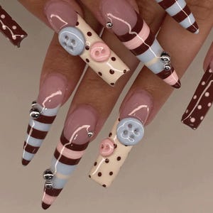 May include: Long, sculpted artificial nails featuring a mix of designs including cream-colored bases with brown polka dots and light blue and pink buttons, alongside striped patterns in brown, light blue, and pink, accented with small silver beads.