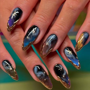 May include: Almond-shaped nail art with a celestial theme. Each nail features a dark base with a gradient of blues, purples, and browns, accented with gold starburst designs and gold trim. The nails are long and well-manicured.