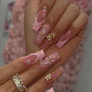 May include: A hand with long, square-shaped, pink acrylic nails. Some nails feature gold floral embellishments and gold outlines. Other nails have a wavy pink design with small, clear rhinestones. A gold ring with clear stones is also visible.