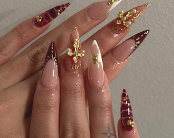May include: A close-up of long, stiletto-shaped artificial nails. The nails feature a variety of designs, including a French manicure with white tips, burgundy and gold accents, and ornate gold embellishments with pearls. Some nails have a textured, reptile-like pattern.