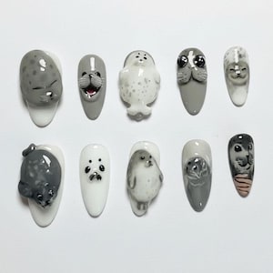 May include: A set of ten artificial nails, each uniquely designed with a 3D seal motif. The nails feature a variety of colors, including gray, white, and black, with detailed seal faces and bodies. The nail shapes are oval and almond.