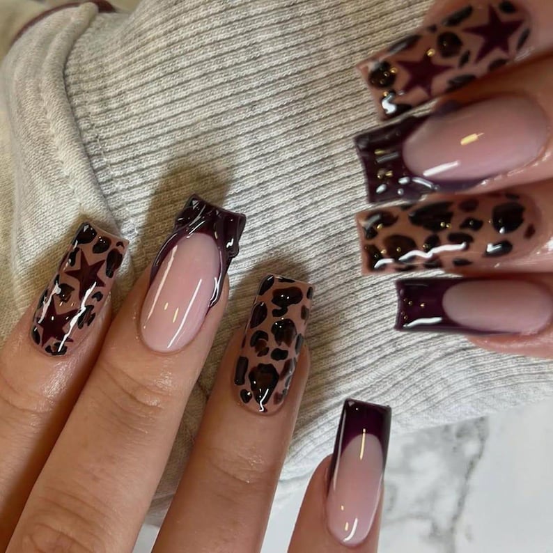 May include: Close-up of manicured nails with diverse designs. Some nails have a nude base with dark burgundy tips, while others display leopard print and star accents. The nails are long and square-shaped, with a glossy finish.