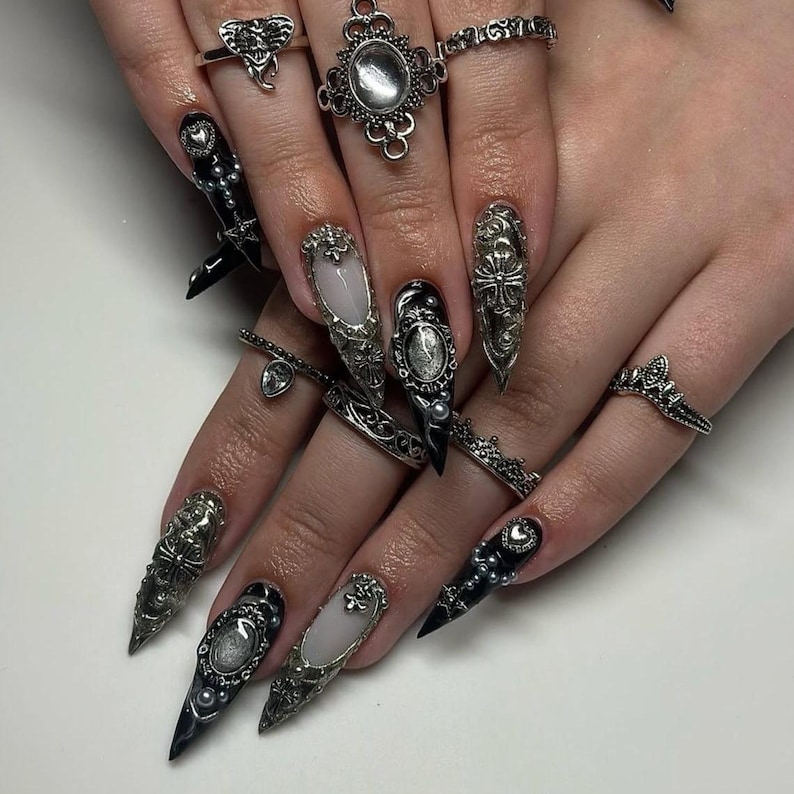 May include: Close-up of hands adorned with elaborate, long, pointed nail art in black, silver, and white. The nails feature intricate designs, including ornate silver embellishments, pearls, and studs. Several silver rings with detailed designs complement the nail art.