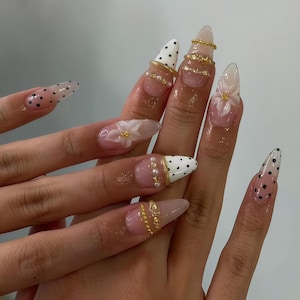 May include: Almond-shaped artificial nails featuring a mix of designs including white tips with black polka dots, clear bases with white 3D floral accents, and gold metallic embellishments including thin bands and small pearl-like beads.