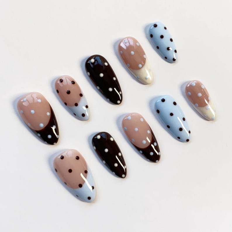 May include: Ten oval false nails in a variety of designs. Some are solid black, others light blue with black polka dots, and some nude with white polka dots and a dark brown tip. The nails are arranged on a white surface.
