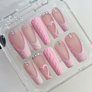 Puede incluir: Un conjunto de u&ntilde;as postizas rosas en una caja de pl&aacute;stico transparente. Las u&ntilde;as presentan una variedad de dise&ntilde;os, incluyendo puntas con purpurina, texturas de punto trenzado y detalles en forma de coraz&oacute;n. Cada u&ntilde;a est&aacute; adornada con peque&ntilde;os brillantes.