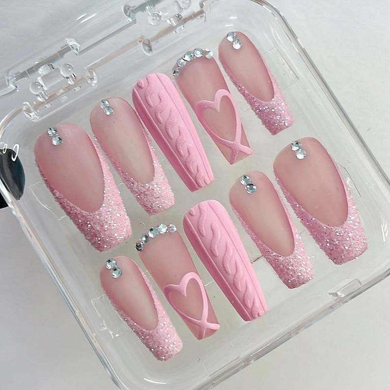 Puede incluir: Un conjunto de u&ntilde;as postizas rosas en una caja de pl&aacute;stico transparente. Las u&ntilde;as presentan una variedad de dise&ntilde;os, incluyendo puntas con purpurina, texturas de punto trenzado y detalles en forma de coraz&oacute;n. Cada u&ntilde;a est&aacute; adornada con peque&ntilde;os brillantes.