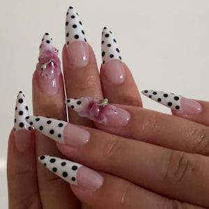 May include: Close-up of a hand with long, pointed acrylic nails. The nails have a white base with black polka dots on the tips. Some nails have a pink base with a floral design. The nails are a fashionable accessory.