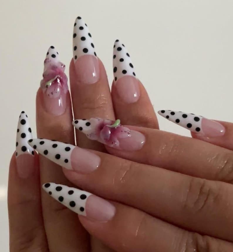 May include: Close-up of a hand with long, pointed acrylic nails. The nails have a white base with black polka dots on the tips. Some nails have a pink base with a floral design. The nails are a fashionable accessory.
