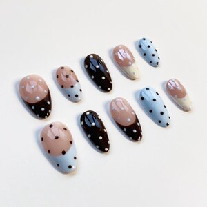 May include: A set of oval false nails in various designs. Some are solid black, others light blue with white polka dots, and some feature light pink and white with black polka dots. The nails are displayed on a white surface.