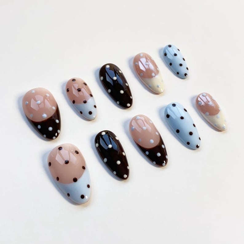 May include: A set of oval false nails in various designs. Some are solid black, others light blue with white polka dots, and some feature light pink and white with black polka dots. The nails are displayed on a white surface.