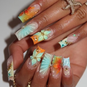 May include: Long square-shaped artificial nails featuring a gradient of orange and light blue. The nails are decorated with three-dimensional sculpted flowers, raised shell patterns, small gold metallic beads, and clear water droplet accents.