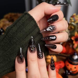 May include: A hand with long, pointed, black and clear acrylic nails. The nails feature various designs, including a spider, a skull hand, and spiral patterns. The nails are decorated with black and silver embellishments, perfect for Halloween.