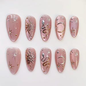 May include: Set of ten press-on nails in a light pink hue, each adorned with celestial designs. The nails feature silver and gold accents, including suns, moons, snakes, and starbursts, creating a mystical aesthetic.