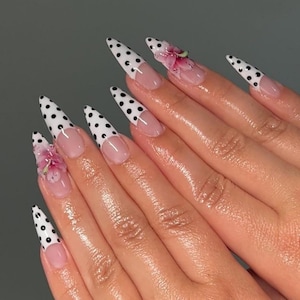 May include: Long, pointed fingernails with a white polka dot design on the tips. Some nails feature a pink floral accent. The nails have a clear base with a pinkish hue, creating a stylish and feminine look.