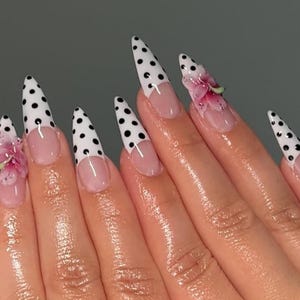 May include: A hand with long, pointed fingernails. The nails are painted with a white base and black polka dots. Some nails have a clear base with a pink flower design. The nails have a French tip design.
