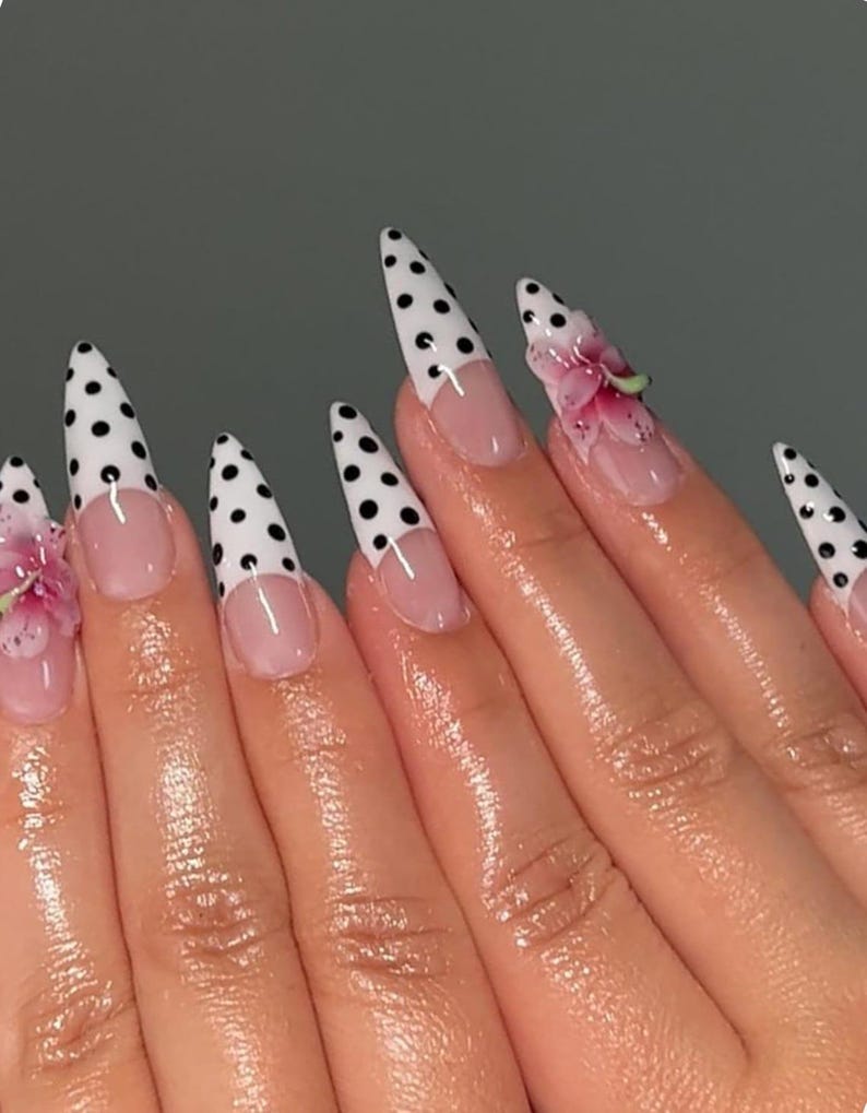 May include: A hand with long, pointed fingernails. The nails are painted with a white base and black polka dots. Some nails have a clear base with a pink flower design. The nails have a French tip design.