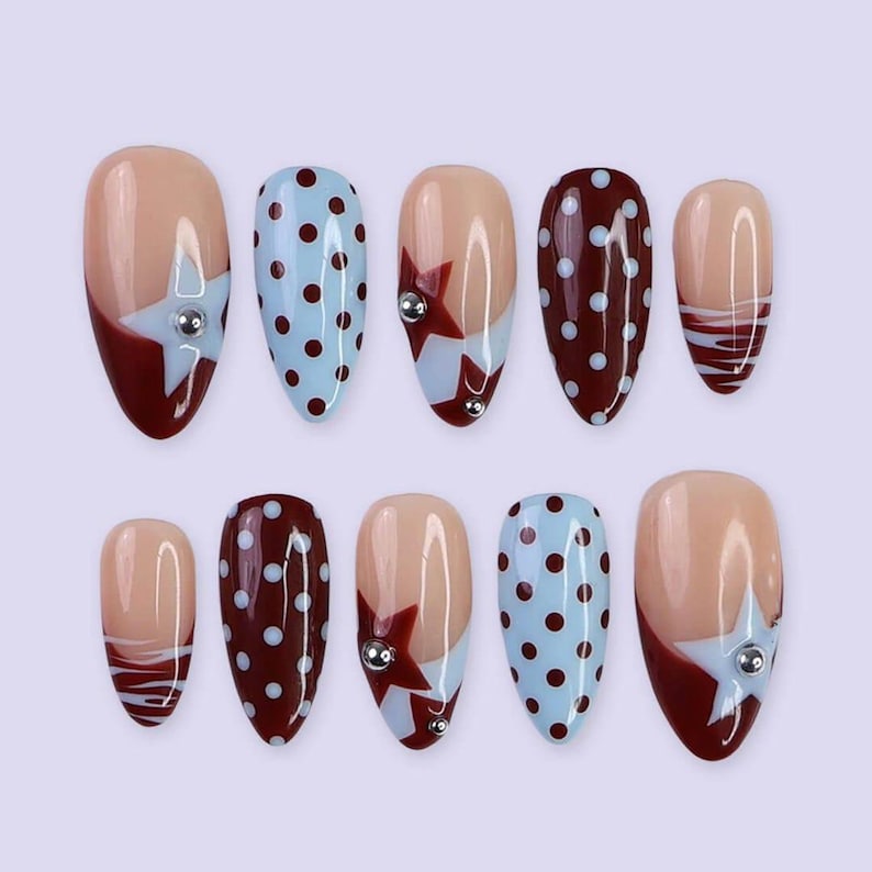 May include: A set of ten almond-shaped false nails. The nails feature a variety of designs, including a nude base with a burgundy tip and a star accent, light blue polka dots, and burgundy polka dots. Some nails have a silver gem.