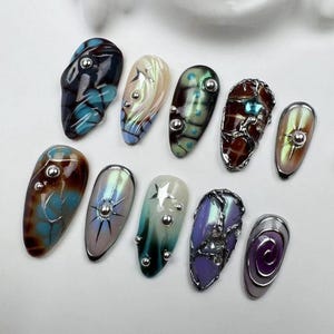 May include: A collection of ten uniquely designed artificial nails. Each nail features a different color scheme and design, including swirls, metallic accents, and embedded silver studs. The nails are oval-shaped and showcase a variety of artistic styles.